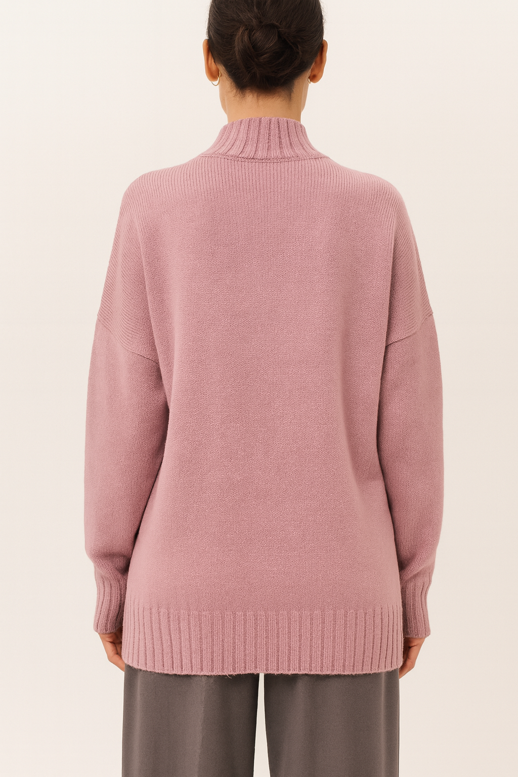 Back view of dusty rose Nordash Oversized Turtleneck Sweater, soft knit with ribbed details, paired with grey trousers in a minimal studio setting.