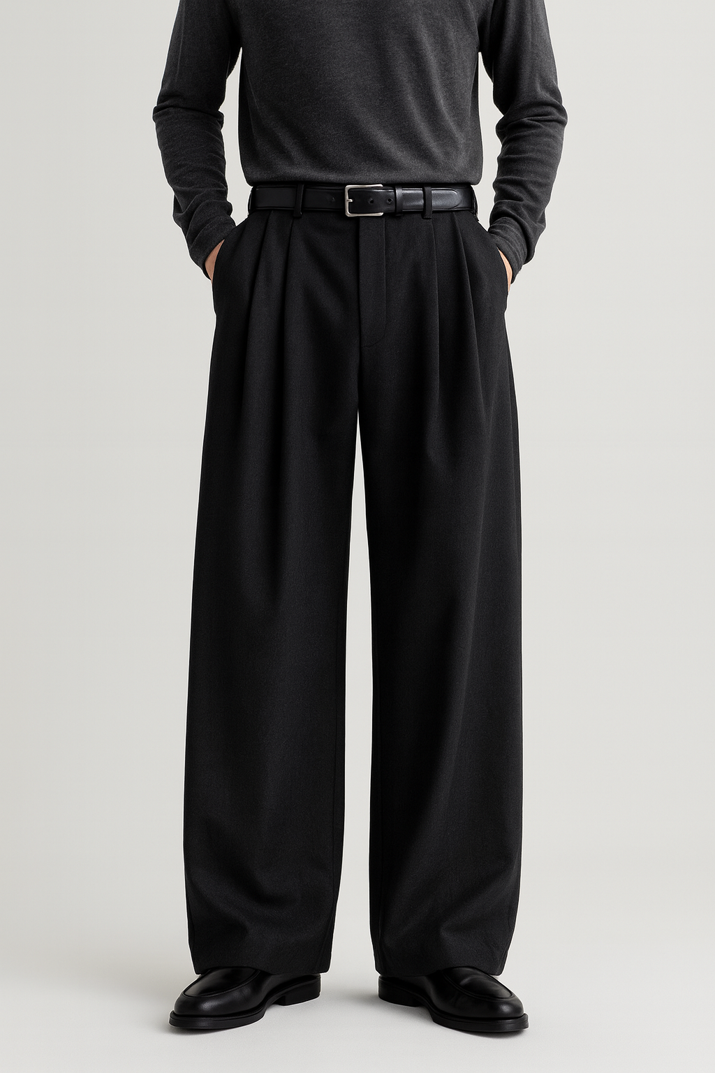 Front view of men’s black wide, full-length pleated trousers with mid-rise waist, deep double pleats, and extra-wide leg, styled with a tucked-in dark grey crewneck shirt, black leather belt, and black loafers against a light-grey background.