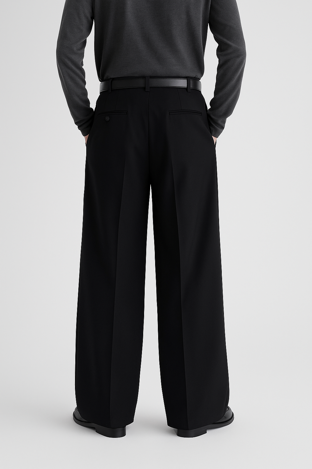 Back view of men’s black wide, full-length pleated trousers with mid-rise waistband, back welt pockets, and extra-wide straight-leg silhouette, styled with a tucked-in dark grey crewneck shirt, black leather belt, and black loafers.